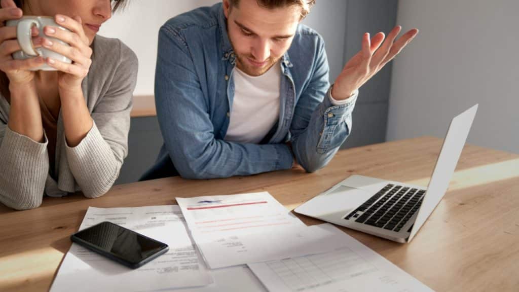 A man looking down at paperwork on a desk with a laptop, gesturing with one hand.