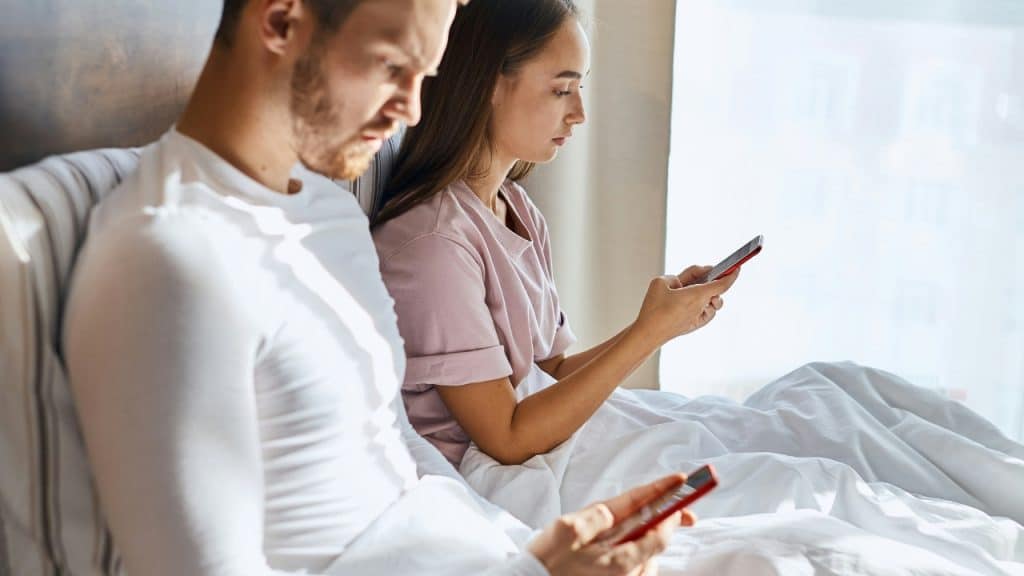 Two people sit side-by-side in bed, both looking down at their individual red mobile phones.