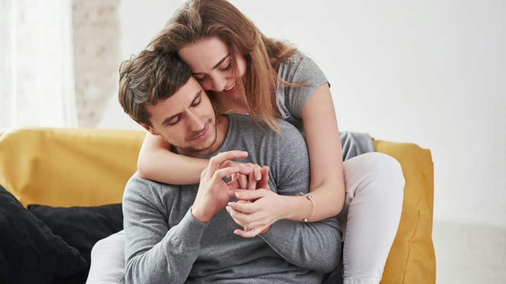 A woman hugs a man from behind while they both look down at their hands.