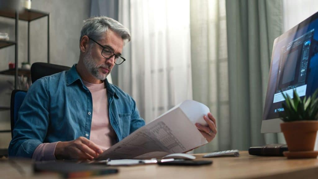 A man with glasses and a beard reviews documents at a desk with a computer.