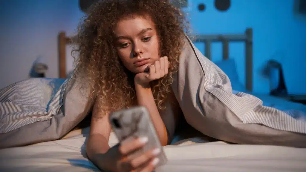 A woman with curly hair lies in bed under a blanket while looking at a smartphone.
