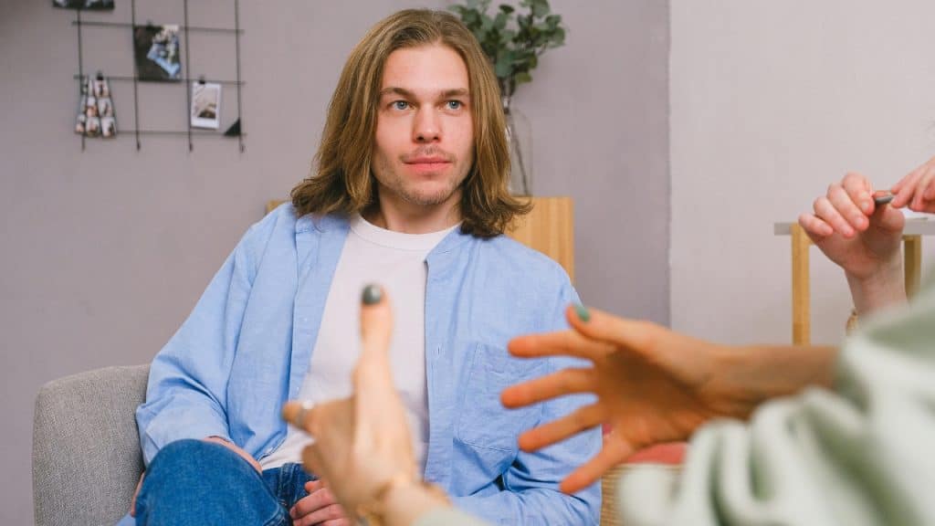 A man with long hair sits on a chair while someone gestures with their hands.