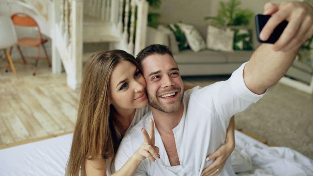 A man and woman smile while taking a selfie together, with the woman gesturing a peace sign.