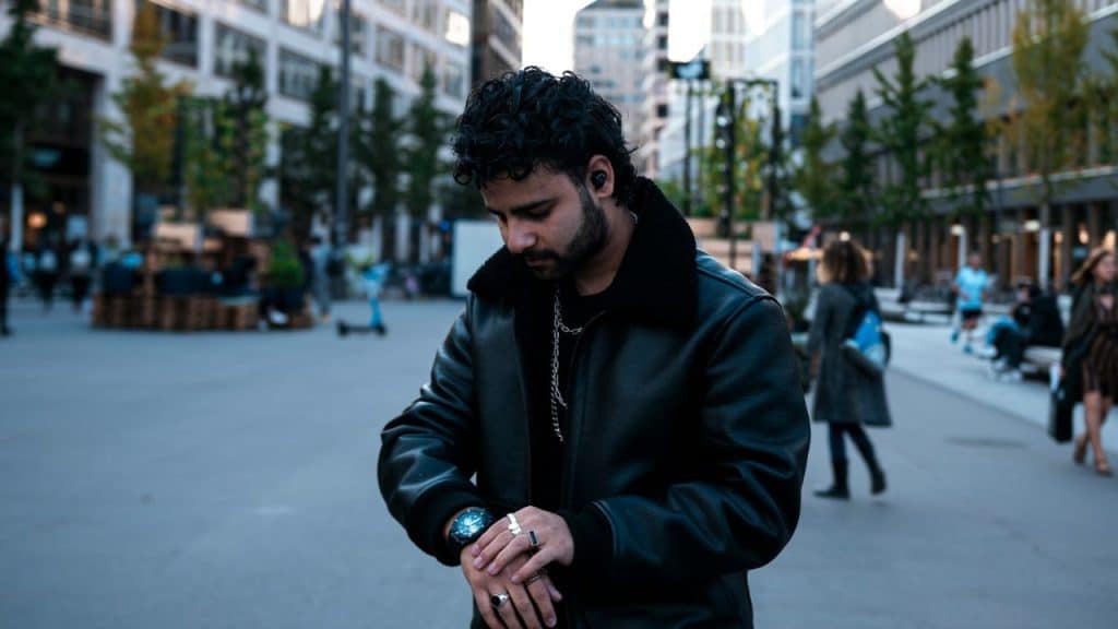 A man in a leather jacket looks down at his watch while standing in a city.