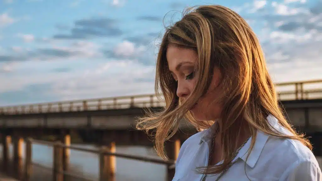 A woman with windblown hair looking down near a waterfront bridge at sunset.