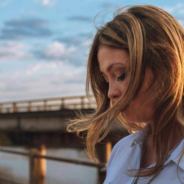 A woman with windblown hair looking down near a waterfront bridge at sunset.