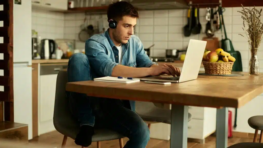 A man wearing headphones works on a laptop at a kitchen table.