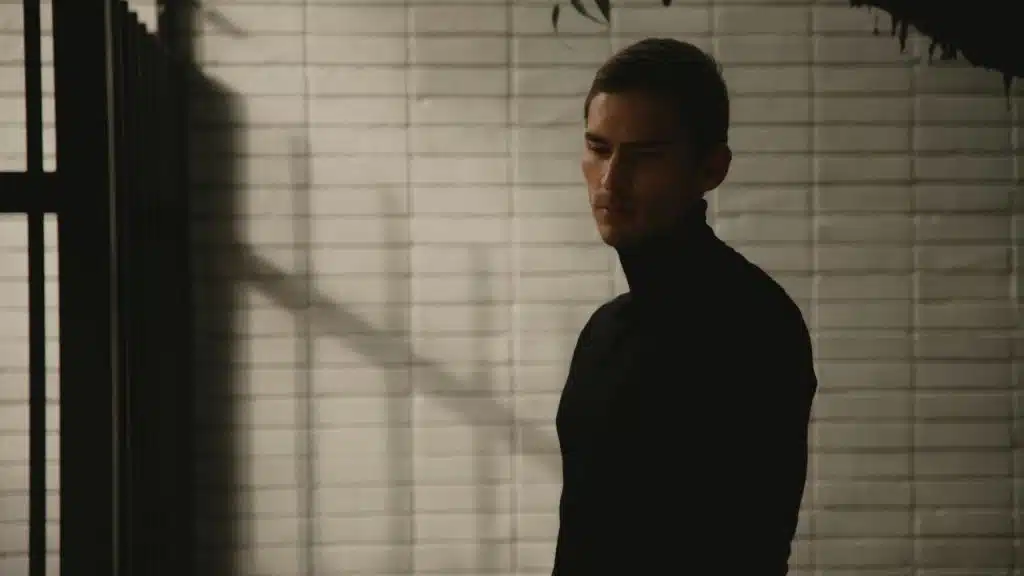 A man in a black turtleneck standing against a tiled wall in soft light.