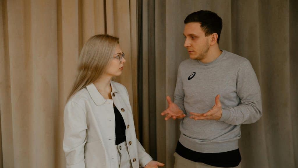 A man with his hands raised speaks to a woman wearing glasses in a room.