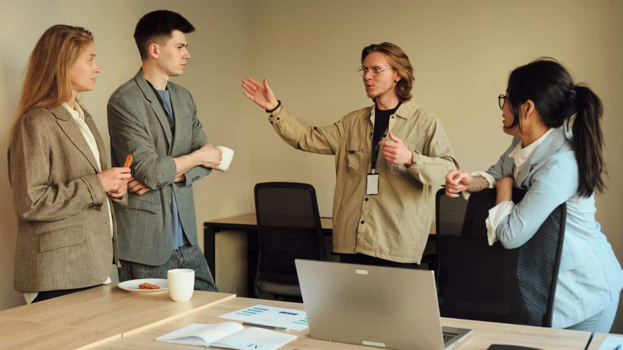 A man gestures and speaks while standing among three colleagues in an office setting.