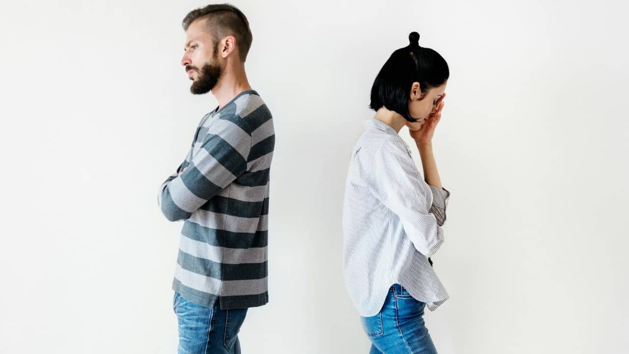A man and woman stand back-to-back; he has arms crossed while she holds her face.