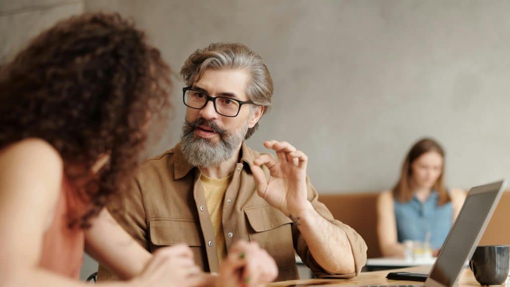 A bearded man with glasses gestures with his hand while talking to a woman at a table.