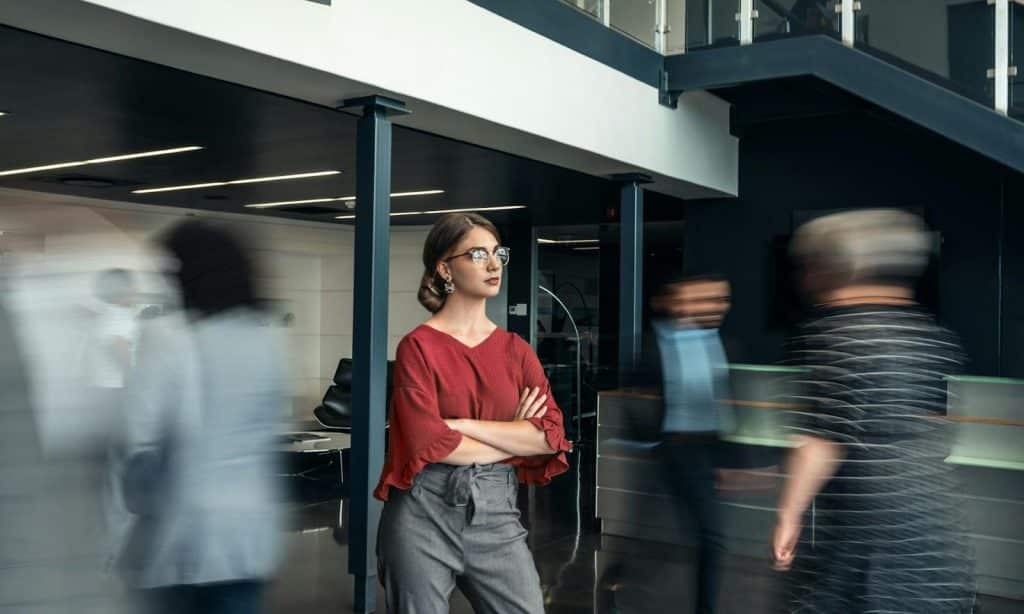 A woman wearing glasses standing in the midst of a crowd in an office with crossed arms