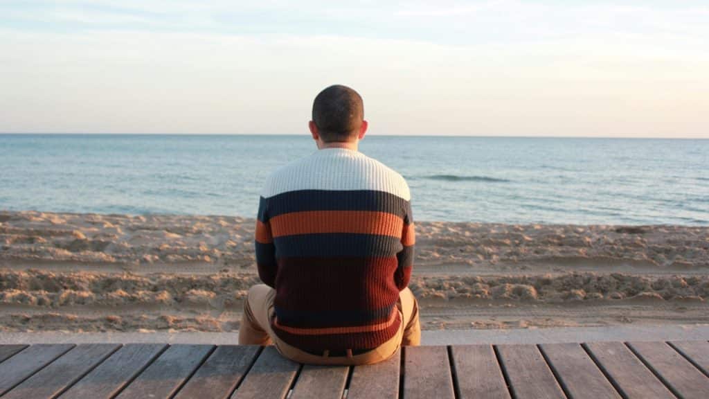 A person viewed from behind sits on a wooden boardwalk facing a calm, blue ocean.