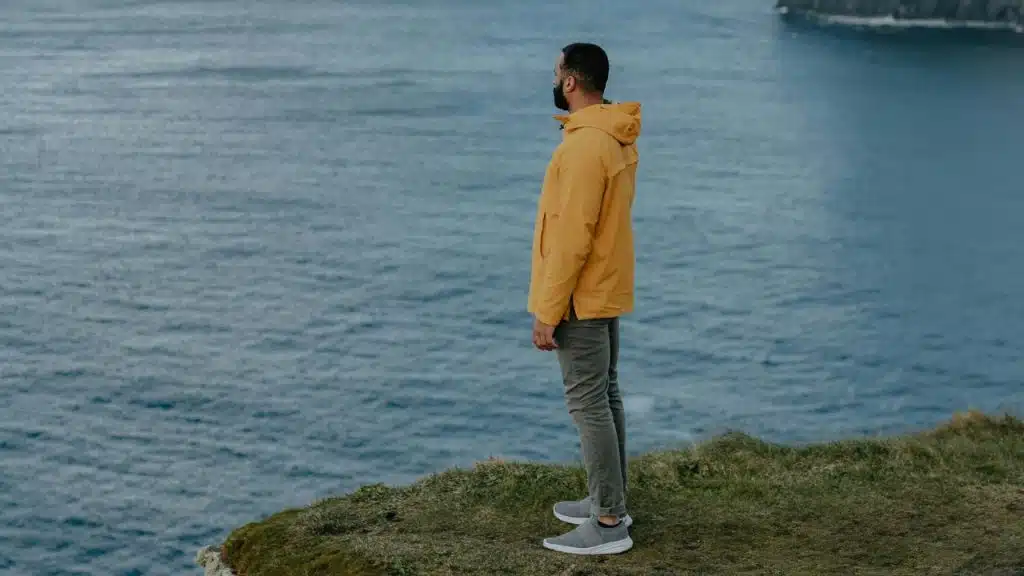 A man in a yellow hooded jacket stands on a grassy cliff overlooking the ocean.