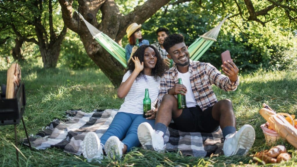 An African American couple sitting in a green garden and taking a selfie on a smartphone while their multiracial friends are talking in a hammock in the background at a summer picnic outdoors.
