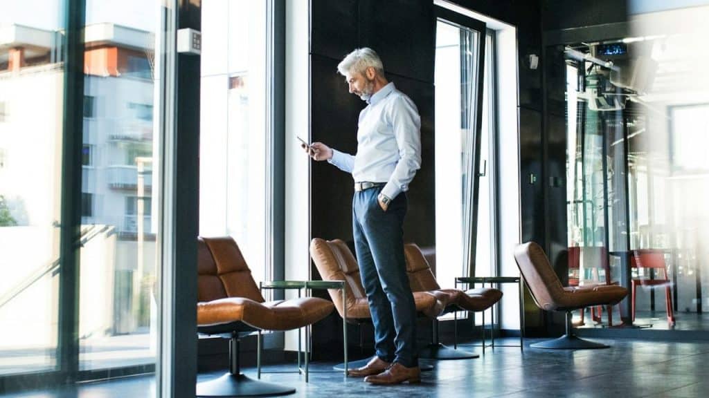 A man in a light blue shirt stands in a modern lounge area looking at his phone.