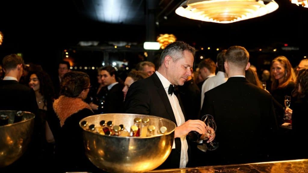 A man in a tuxedo pours a drink at a crowded, dimly lit formal event.