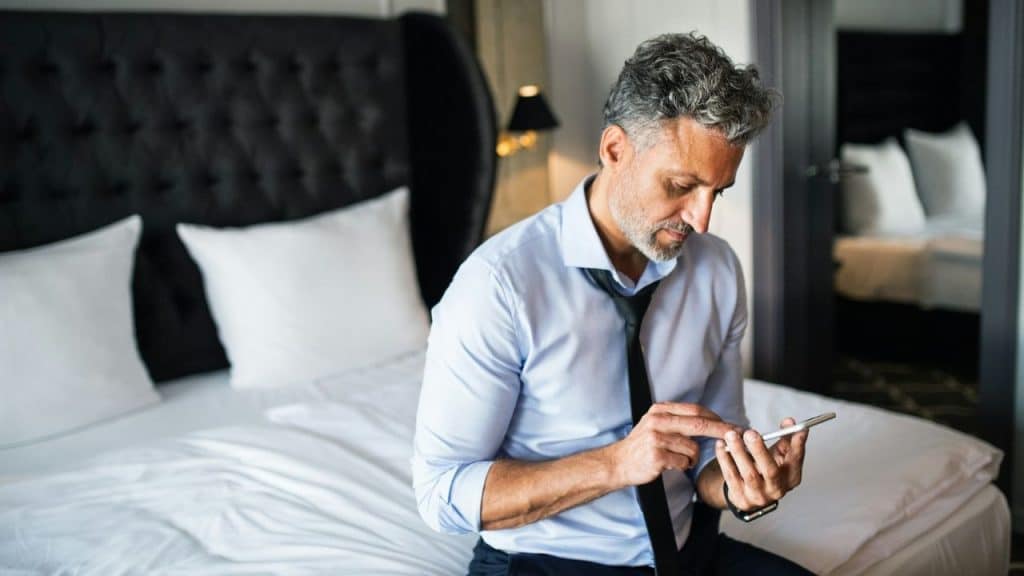 A man in a dress shirt and loosened tie sits on a bed using a smartphone.