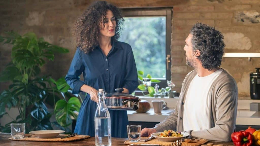 A woman brings a pot to a wooden dining table where a man is sitting.