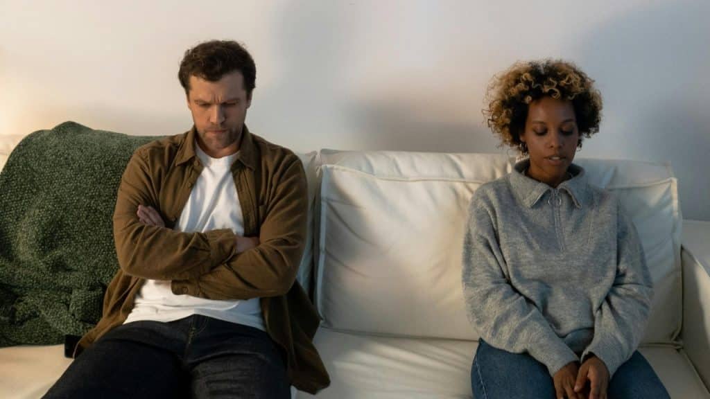 A man and woman sit on a white sofa with space between them, looking away.