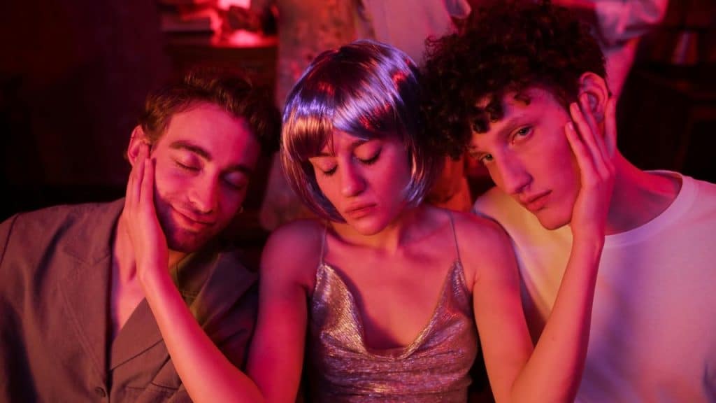 Three young people posing closely together under vibrant pink and purple neon club lighting.