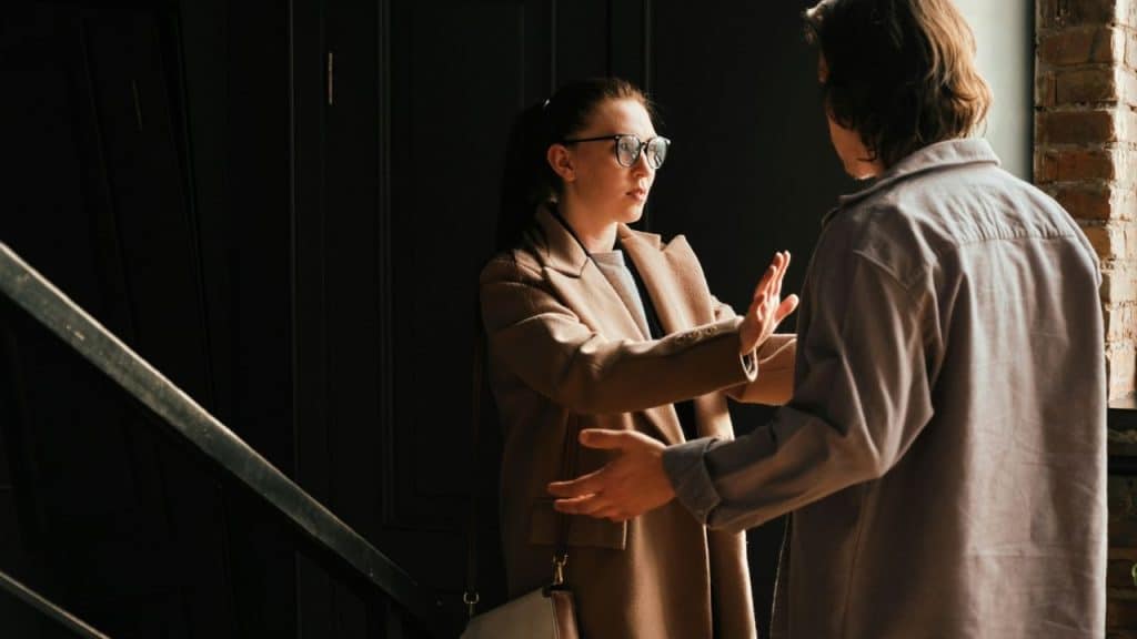 A woman in a tan coat holds her hands up toward a man in a doorway.