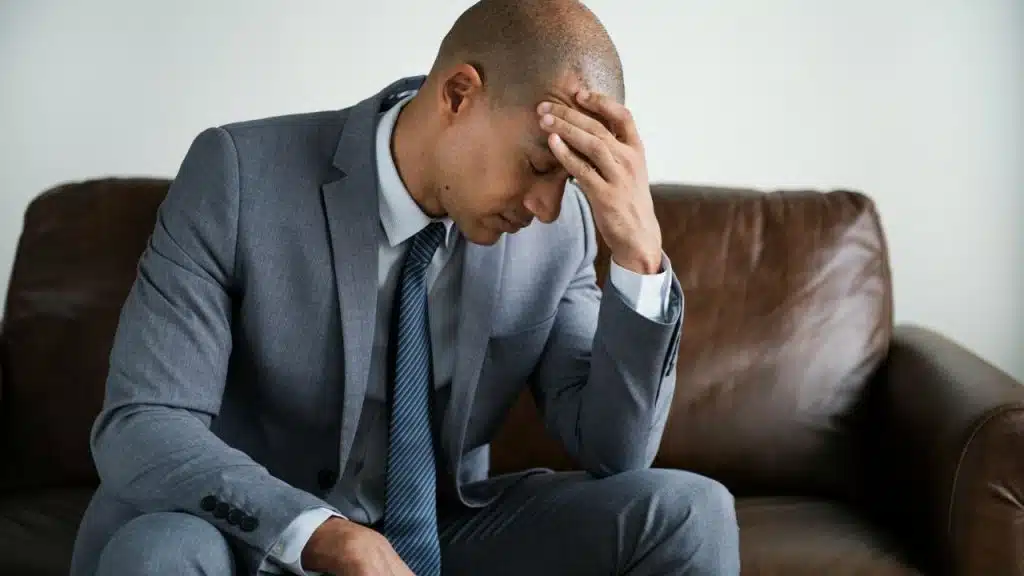 A man in a grey suit sits on a leather sofa with his hand on his forehead.