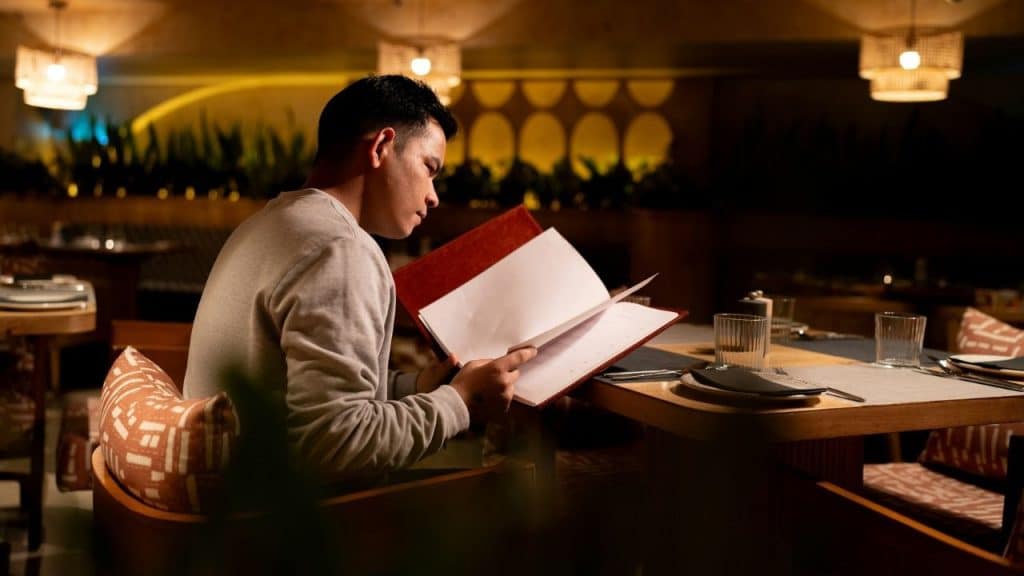 A man sits alone at a restaurant table, carefully reading a large open menu.