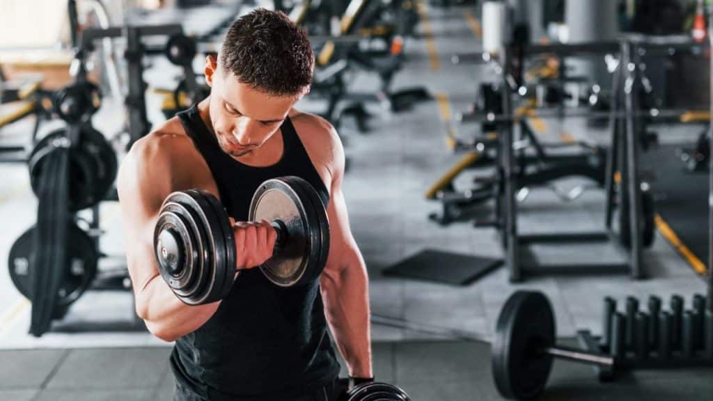 A muscular man in a black tank top lifts a heavy dumbbell in a gym.