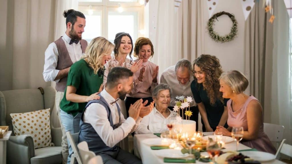 A multi-generational family gathers around a table, clapping as an elderly woman sits before a cake.