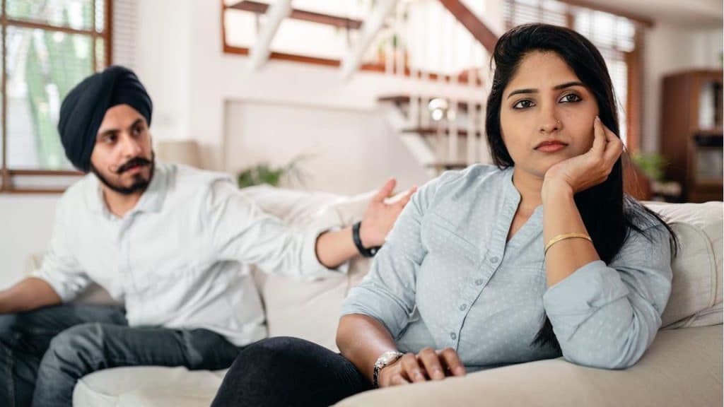 A woman is sitting on a couch with a distant expression while a man in a turban sits behind her, gesturing toward her