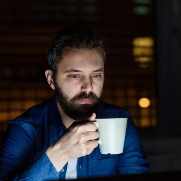 A man with a beard sits in the dark, holding a mug and looking at a screen.