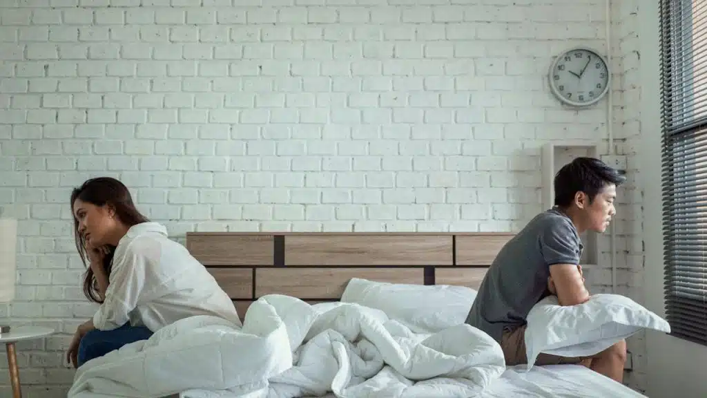A couple sitting on separate ends of a bed facing away from each other.