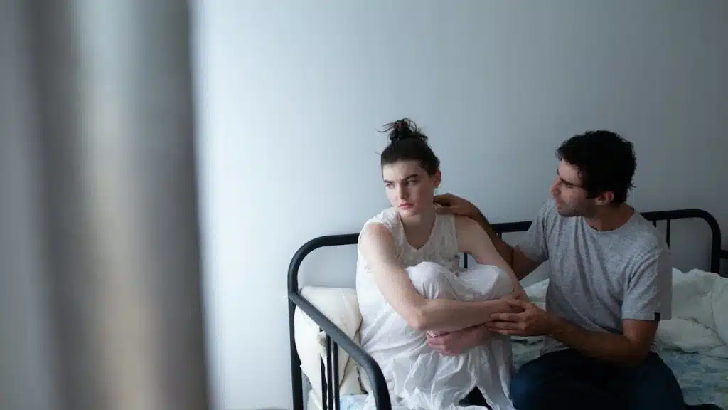 A man gently touches the shoulder of a sad or troubled woman sitting curled up on a metal bed frame