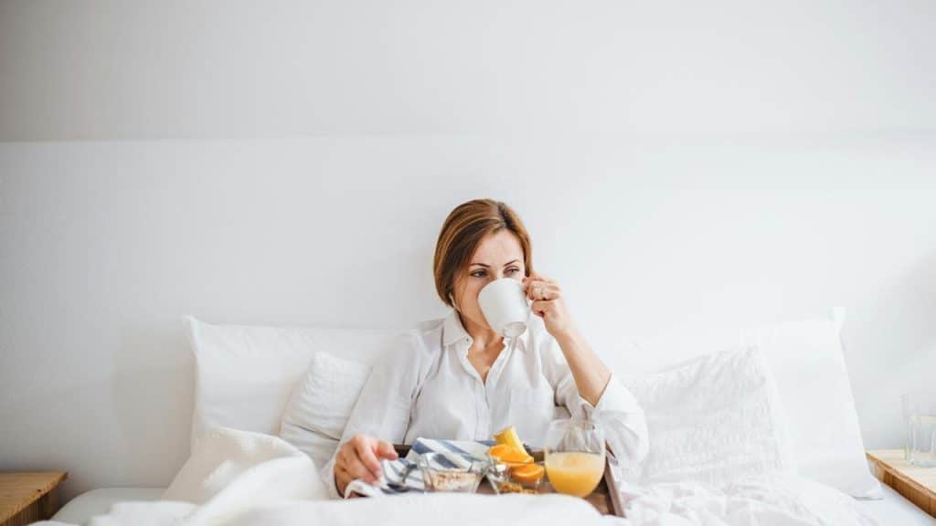 A woman in bed drinking coffee with breakfast on a tray.