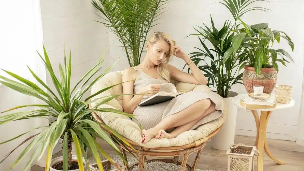A woman sitting in a chair surrounded by plants while reading a book.