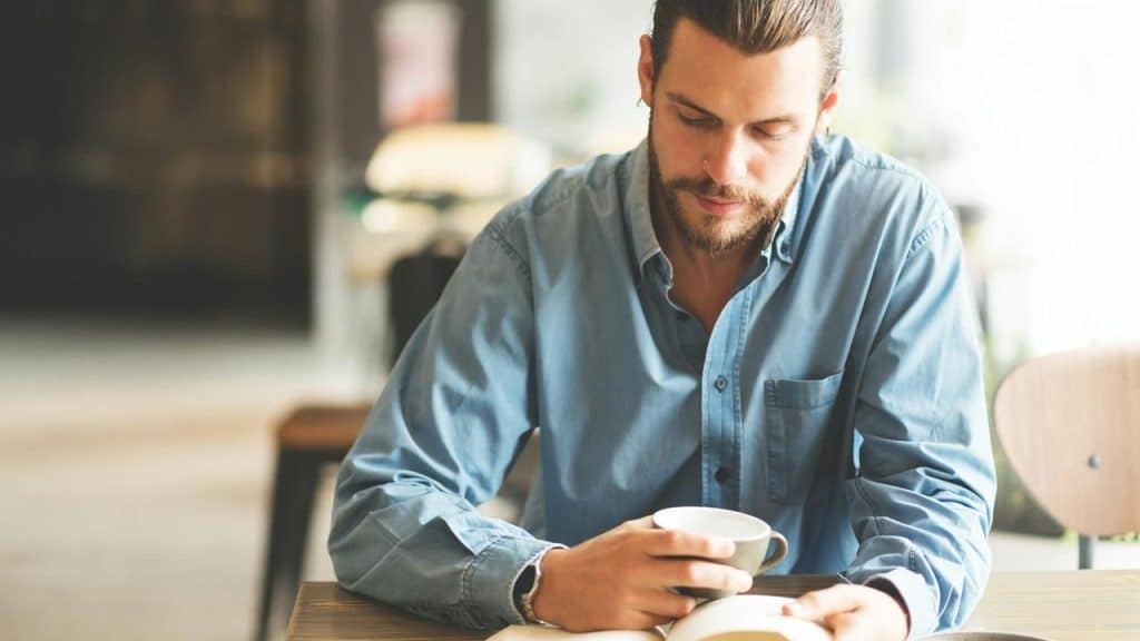 A man is reading a book while holding a cup of coffee.