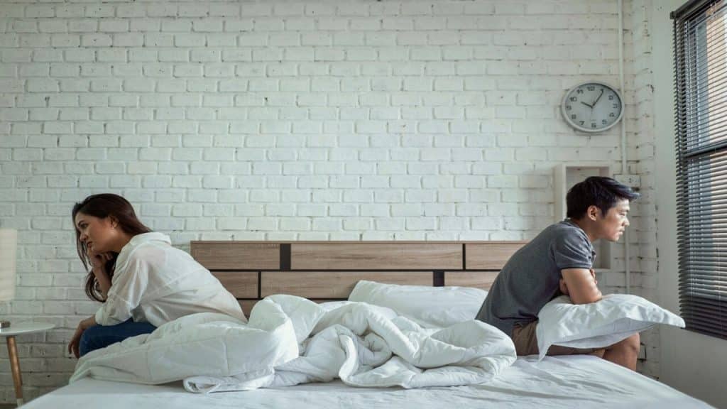 A couple sitting on opposite sides of a bed with their backs against each other.