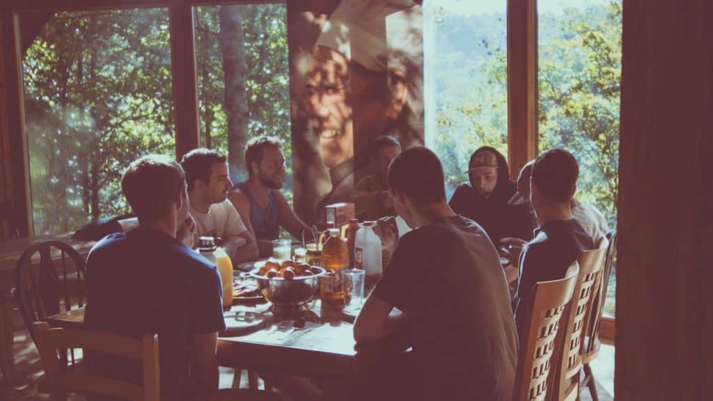 A group of men sharing a meal together at a large wooden table in a room with large windows overlooking a forest.