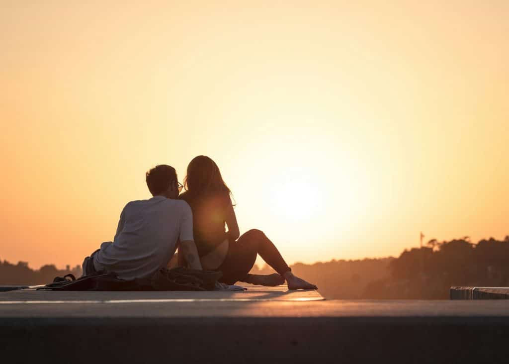 A couple sitting on a ledge watching the sunset together.
