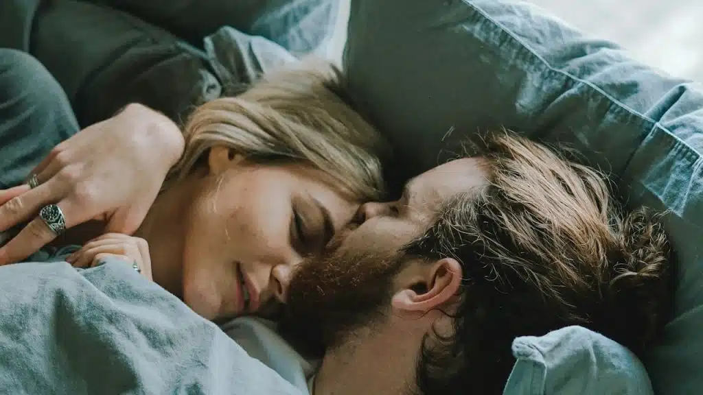 A bearded woman kissing a woman’s forehead while they lie on a bed. 