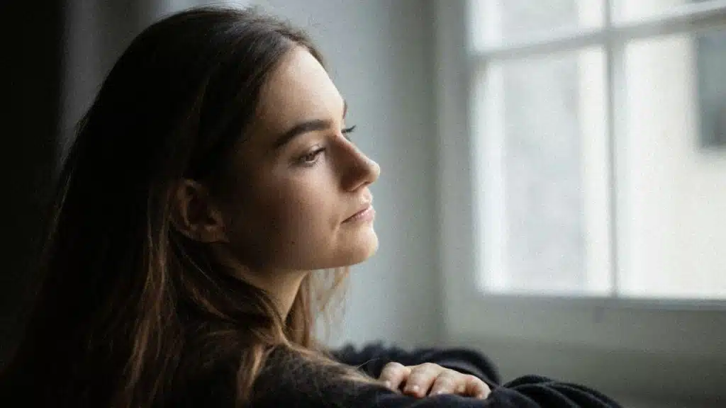 Woman with long brown hair looking out a bright window with a thoughtful expression.