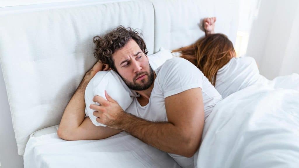 A man is lying in bed looking worried while a woman sleeps next to him with her back turned