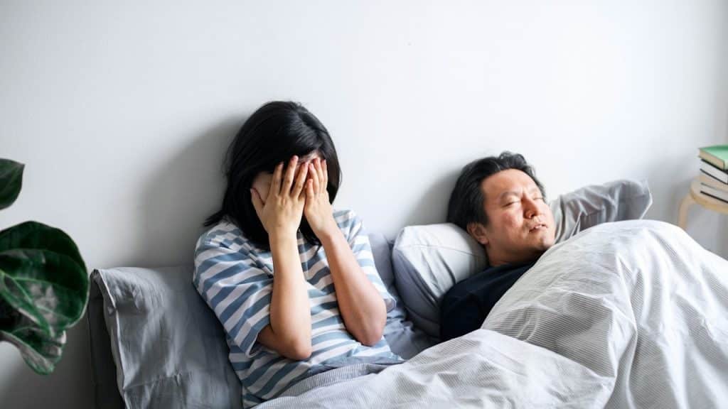 A stressed woman with hands on face siitin in bed with a man sleeping beside her.