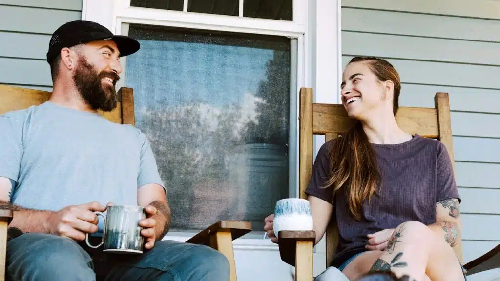 A cheerful couple sitting in wooden rocking chairs on a porch, laughing and holding coffee mugs