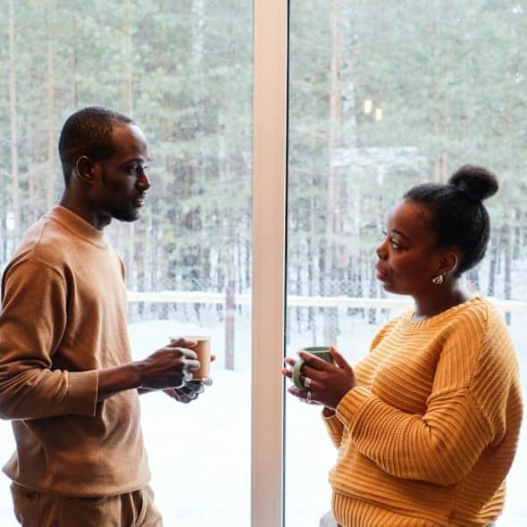 A couple talking and having coffee by a window outside of which there are trees and snow covered ground