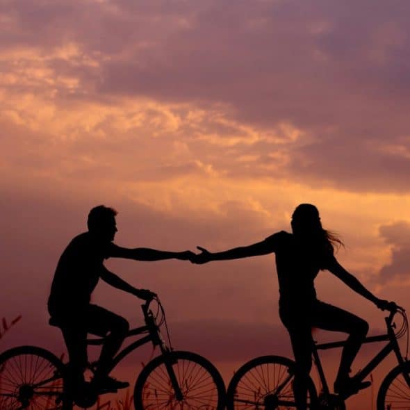 A woman on bike reaching for the man's hand behind her also on the bike.