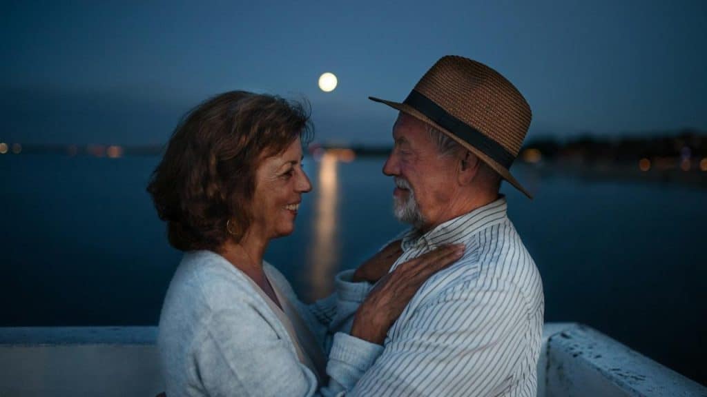 A happy mature couple hugging outdoors on a pier by the sea at dusk, looking at each other.