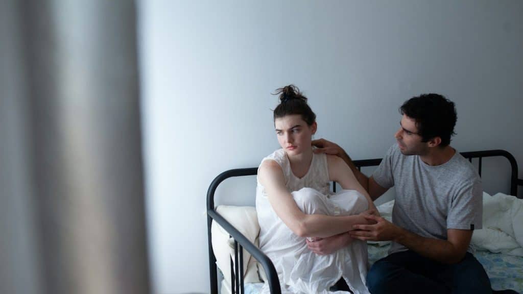 A man gently touches the shoulder of a sad or troubled woman sitting curled up on a metal bed frame.